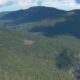 Aerial view of a dense evergreen forest covering rolling mountain ridges under a partly cloudy sky.