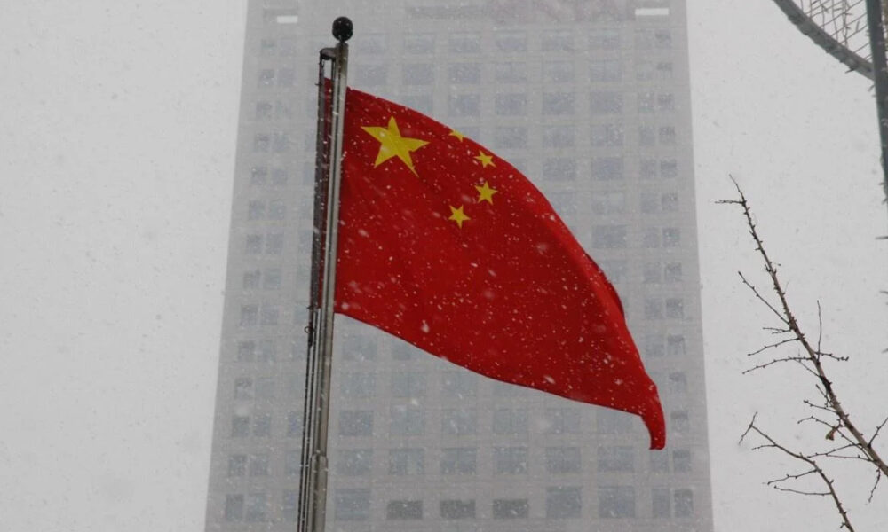 Red Chinese national flag fluttering on a flagpole in a snowy urban scene with a tall building in the background.