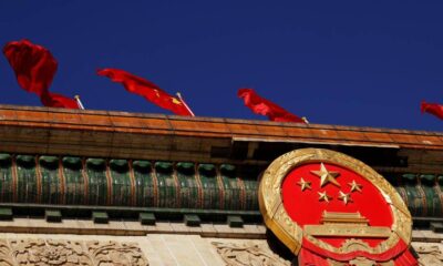 Close-up of China's national emblem mounted on a building, with red flags fluttering against a deep blue sky.