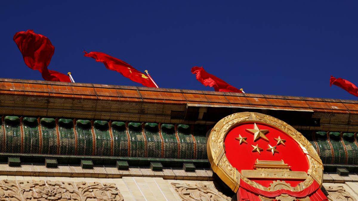 Close-up of China's national emblem mounted on a building, with red flags fluttering against a deep blue sky.