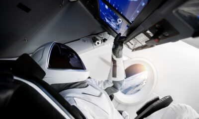 Astronaut in white suit and helmet reaches for cockpit controls inside a spacecraft, Earth visible through a circular window behind.