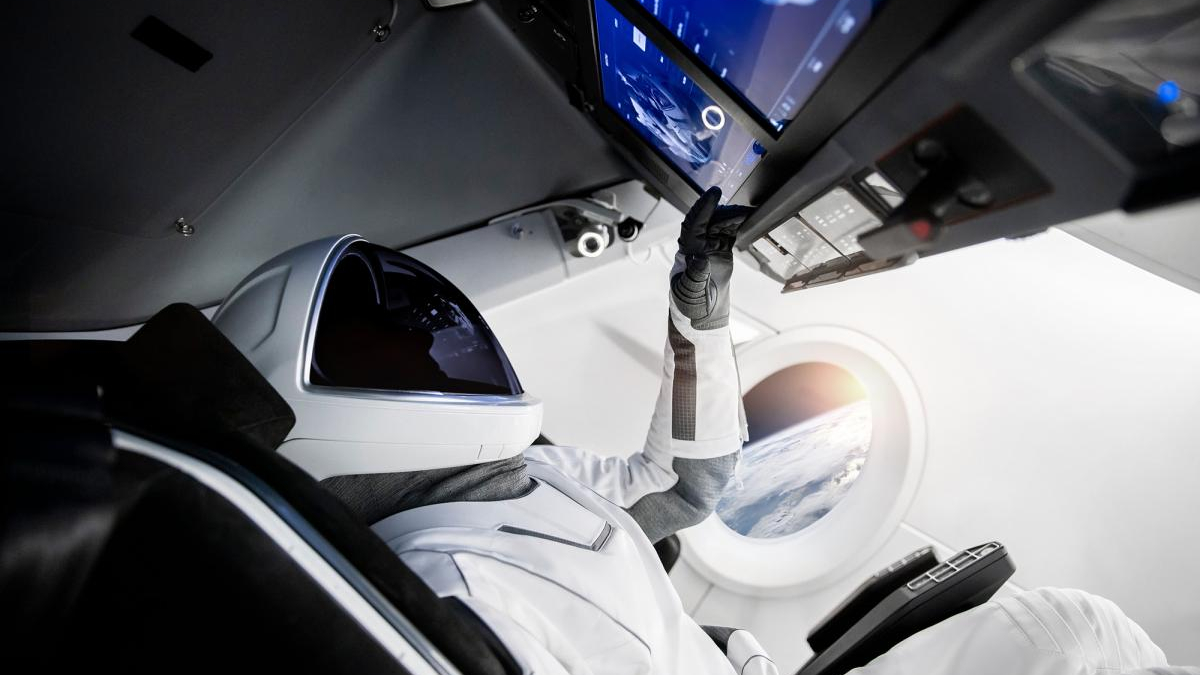 Astronaut in white suit and helmet reaches for cockpit controls inside a spacecraft, Earth visible through a circular window behind.