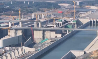 Large-scale bridge construction over a canal with yellow cranes, concrete supports, and unfinished roadway sections; waterway on both sides surrounded by concrete barriers