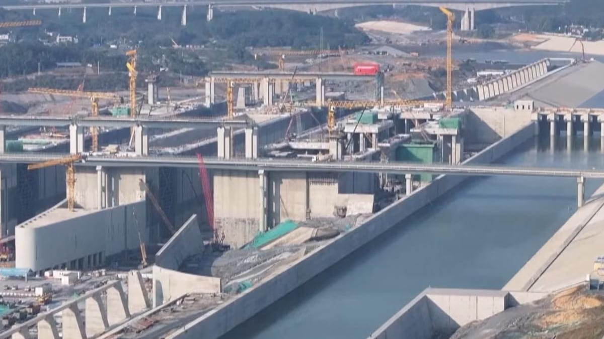 Large-scale bridge construction over a canal with yellow cranes, concrete supports, and unfinished roadway sections; waterway on both sides surrounded by concrete barriers