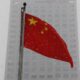 Chinese flag waving on a flagpole in snowy urban setting, with a tall building in the background.