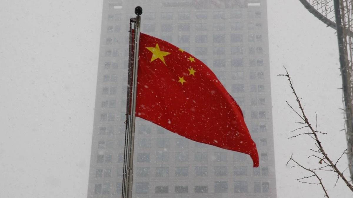 Chinese flag waving on a flagpole in snowy urban setting, with a tall building in the background.