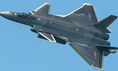 F-22 Raptor stealth fighter jet in flight against a clear blue sky, viewed from below with dark gray camouflage and twin vertical tails nearby.