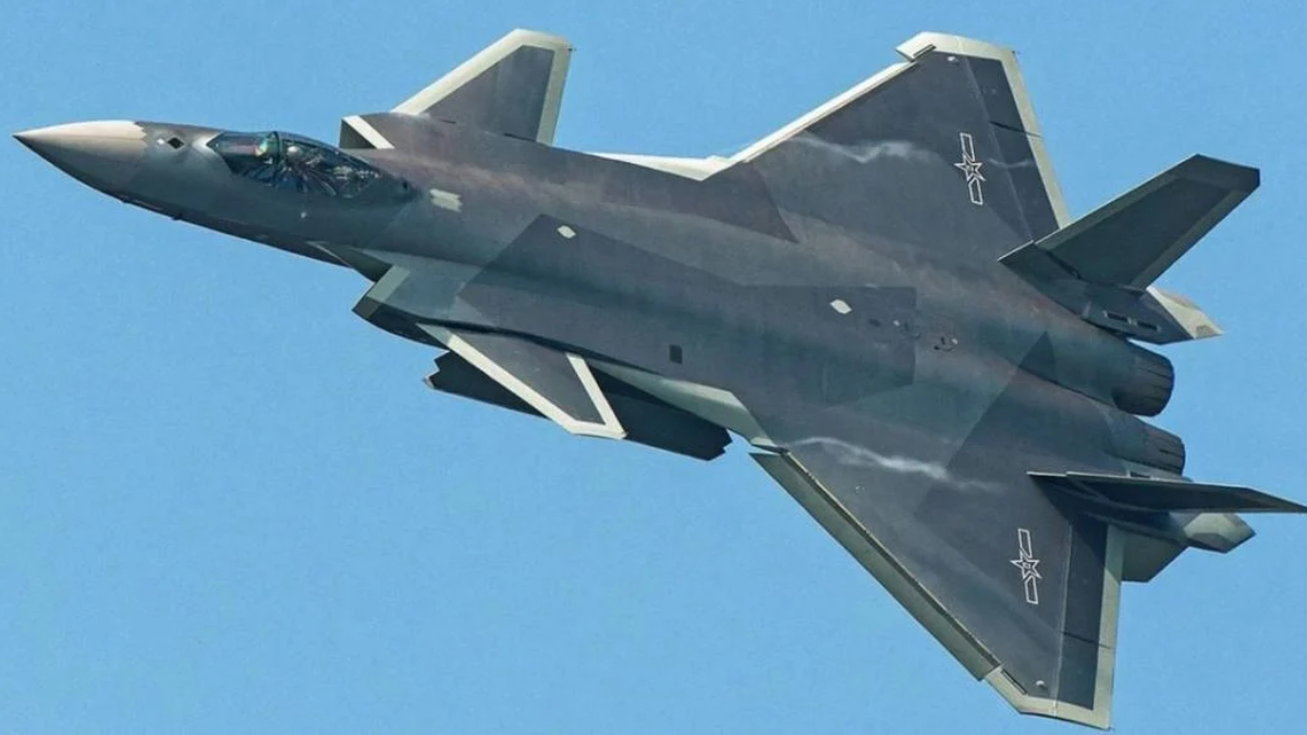 F-22 Raptor stealth fighter jet in flight against a clear blue sky, viewed from below with dark gray camouflage and twin vertical tails nearby.