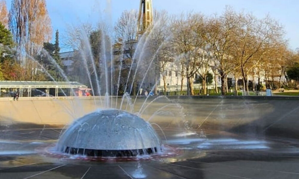 Round fountain with a metal dome in a circular pool, water jets shooting outward in a park setting with trees in the background.