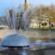 Round fountain with a metal dome in a circular pool, water jets shooting outward in a park setting with trees in the background.
