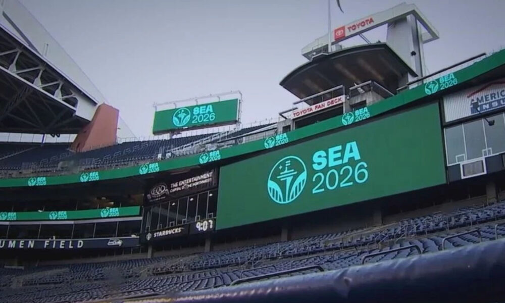 Stadium concourse with a large LED screen reading SEA 2026 and a teal logo, with Lumen Field signage visible in the stands.