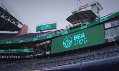 Stadium concourse with a large LED screen reading SEA 2026 and a teal logo, with Lumen Field signage visible in the stands.