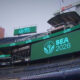 Stadium concourse with a large LED screen reading SEA 2026 and a teal logo, with Lumen Field signage visible in the stands.
