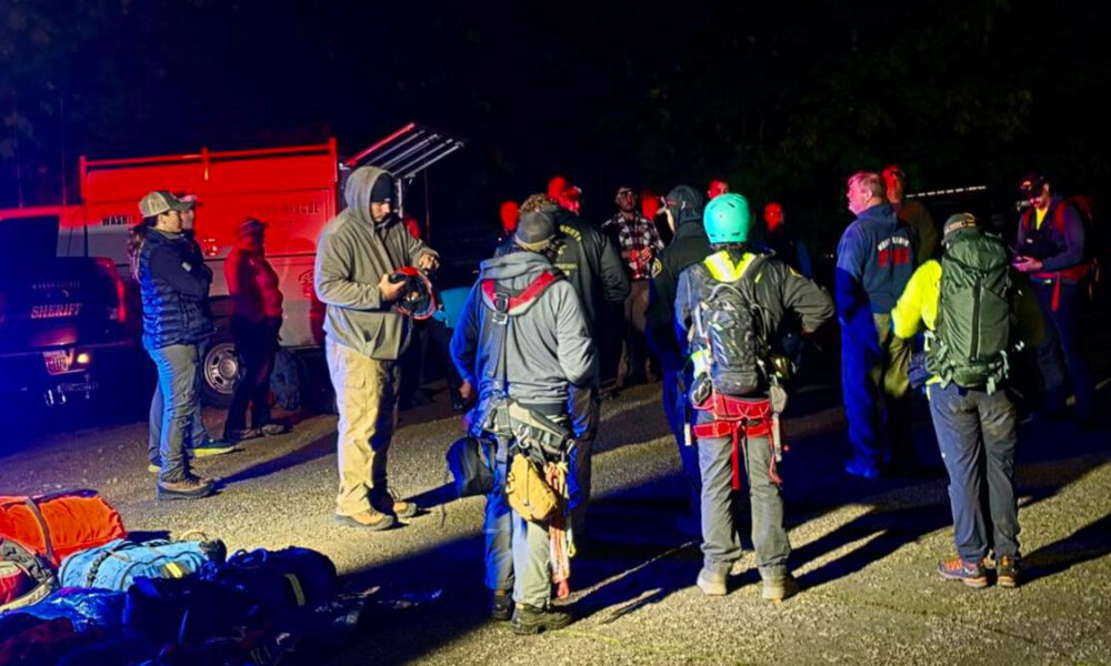Nighttime rescue briefing: group of climbers with helmets and harnesses gathered on an asphalt lot, rescue gear and bags spread out, emergency vehicles in background