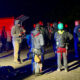 Nighttime rescue briefing: group of climbers with helmets and harnesses gathered on an asphalt lot, rescue gear and bags spread out, emergency vehicles in background