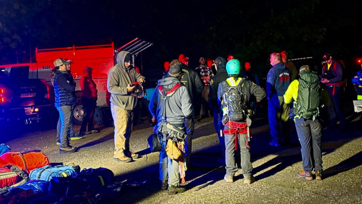 Nighttime rescue briefing: group of climbers with helmets and harnesses gathered on an asphalt lot, rescue gear and bags spread out, emergency vehicles in background