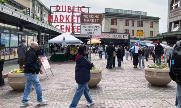 People walk and browse stalls at a busy outdoor farmers market plaza under vintage storefront signs.