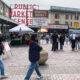 People walk and browse stalls at a busy outdoor farmers market plaza under vintage storefront signs.