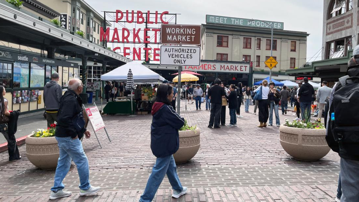 People walk and browse stalls at a busy outdoor farmers market plaza under vintage storefront signs.