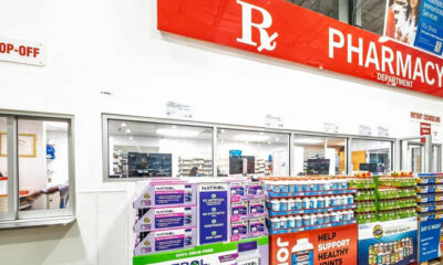 Wide shot of a pharmacy interior with a large red 'PHARMACY' sign above, checkout windows, and shelves stocked with vitamins and supplements in the foreground.