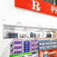 Wide shot of a pharmacy interior with a large red 'PHARMACY' sign above, checkout windows, and shelves stocked with vitamins and supplements in the foreground.
