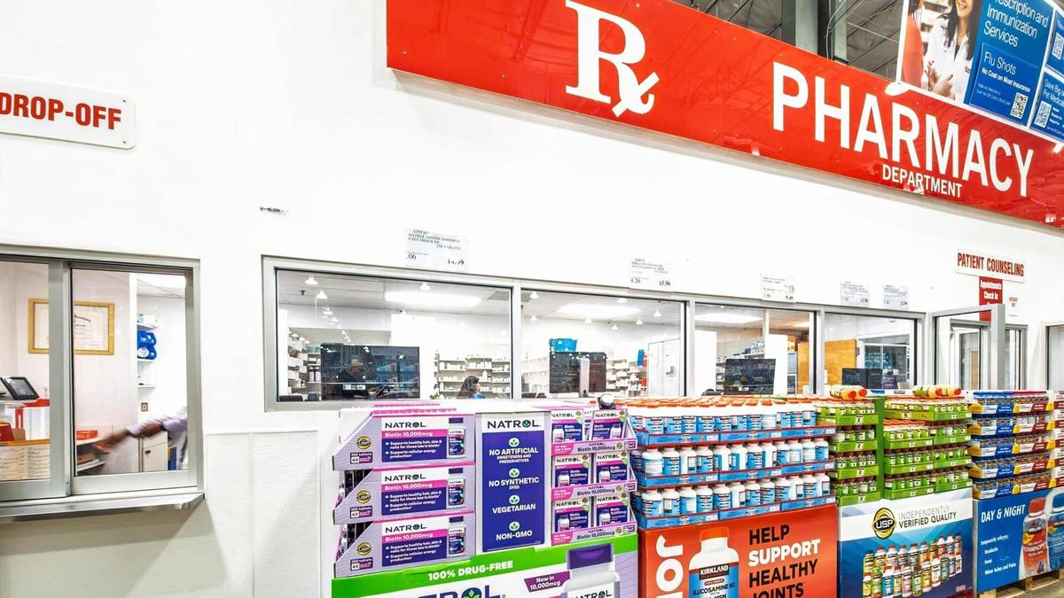 Wide shot of a pharmacy interior with a large red 'PHARMACY' sign above, checkout windows, and shelves stocked with vitamins and supplements in the foreground.