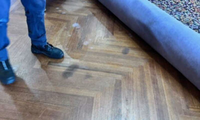 Person in blue pants and black boots standing on a wooden parquet floor with a rolled-up purple carpet partially unrolled nearby, suggesting carpet installation or removal.