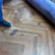 Person in blue pants and black boots standing on a wooden parquet floor with a rolled-up purple carpet partially unrolled nearby, suggesting carpet installation or removal.