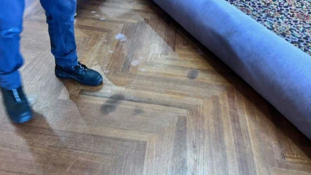 Person in blue pants and black boots standing on a wooden parquet floor with a rolled-up purple carpet partially unrolled nearby, suggesting carpet installation or removal.