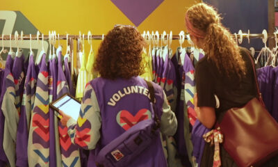 Two volunteers in purple jackets organize colorful jackets on a rack, one checking a tablet while a shopper looks on.