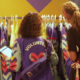 Two volunteers in purple jackets organize colorful jackets on a rack, one checking a tablet while a shopper looks on.