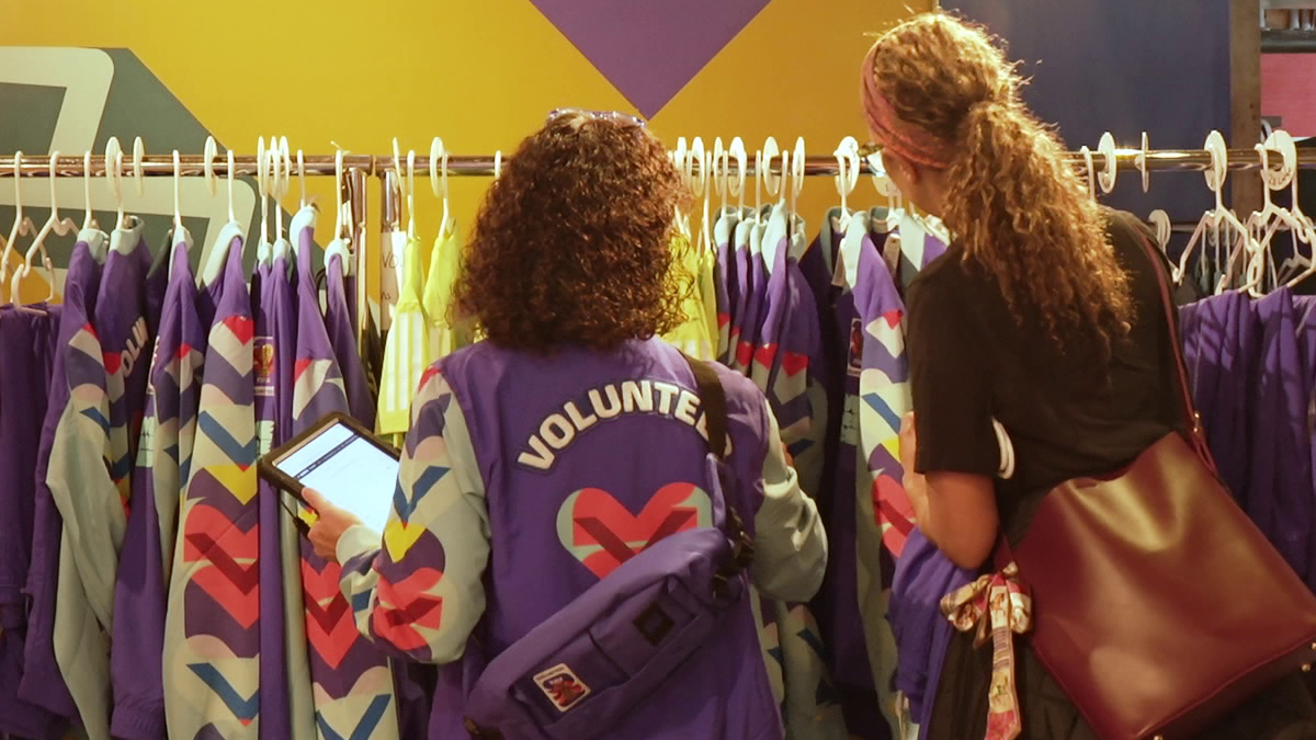 Two volunteers in purple jackets organize colorful jackets on a rack, one checking a tablet while a shopper looks on.