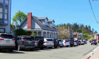 Residential street with a row of parked SUVs and cars beside a blue-grey house with a porch and chimney, sunny day.
