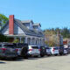 Residential street with a row of parked SUVs and cars beside a blue-grey house with a porch and chimney, sunny day.
