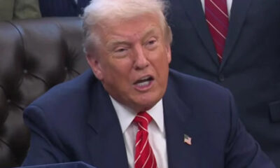Man in a navy suit and red striped tie speaks, wearing an American flag pin, seated on a brown leather couch in a formal setting.