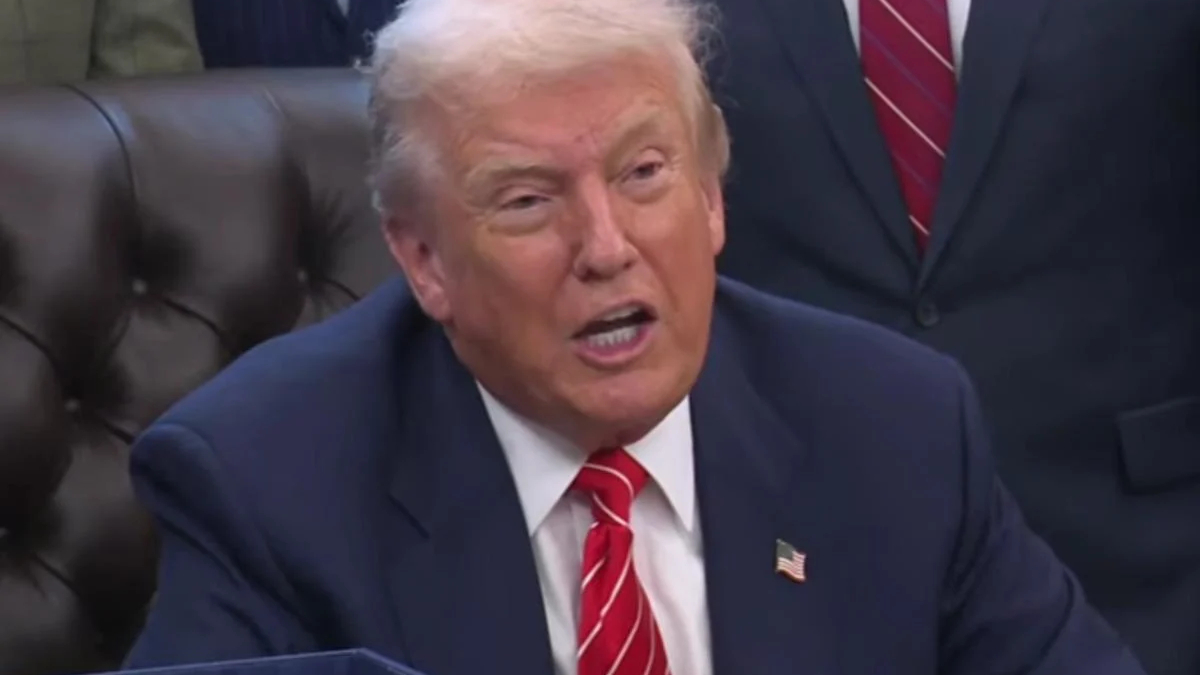 Man in a navy suit and red striped tie speaks, wearing an American flag pin, seated on a brown leather couch in a formal setting.