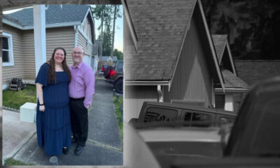 Smiling couple standing together in a driveway outside a beige house; woman in a blue dress, man in a purple shirt and dark pants.