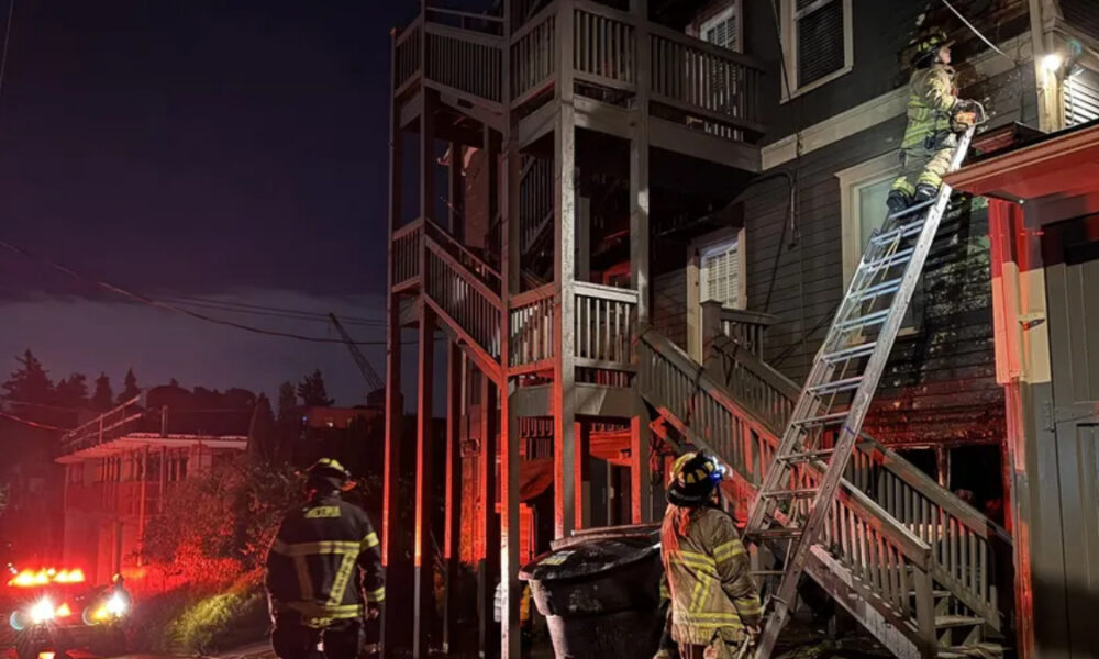 Firefighters at night work a ladder against a two-story house during a nighttime fire response with emergency lights visible.