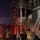 Firefighters at night work a ladder against a two-story house during a nighttime fire response with emergency lights visible.