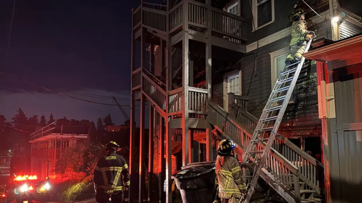 Firefighters at night work a ladder against a two-story house during a nighttime fire response with emergency lights visible.