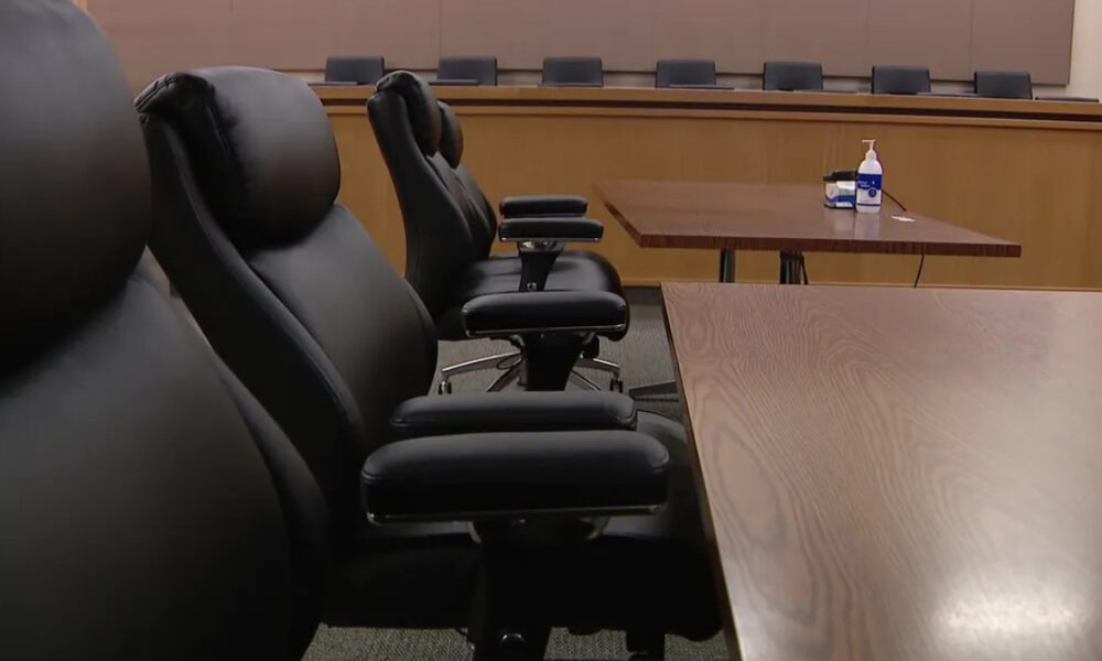 Empty conference room with black leather chairs around a long wooden table; hand sanitizer and tissues on a side table in the background.