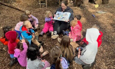 Teacher reads a story to a circle of preschool children outdoors on wood chips in a wooded area.