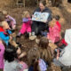 Teacher reads a story to a circle of preschool children outdoors on wood chips in a wooded area.