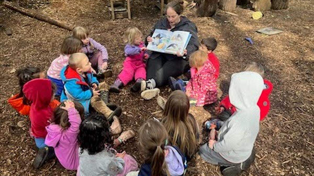 Teacher reads a story to a circle of preschool children outdoors on wood chips in a wooded area.