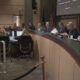 King County Council chamber with officials seated at a curved dais, an American flag, and a large screen in the background.
