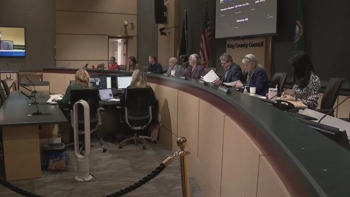 King County Council chamber with officials seated at a curved dais, an American flag, and a large screen in the background.