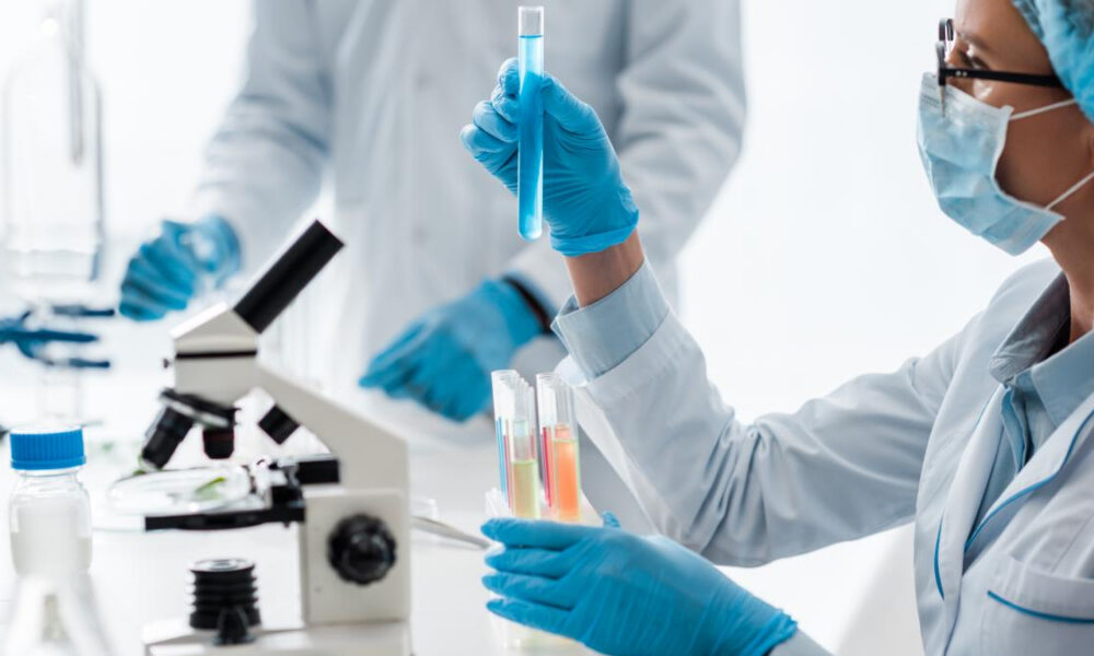 Lab technician in gloves and a mask holds a test tube with blue liquid, with a microscope and more tubes in the background.
