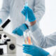 Lab technician in gloves and a mask holds a test tube with blue liquid, with a microscope and more tubes in the background.
