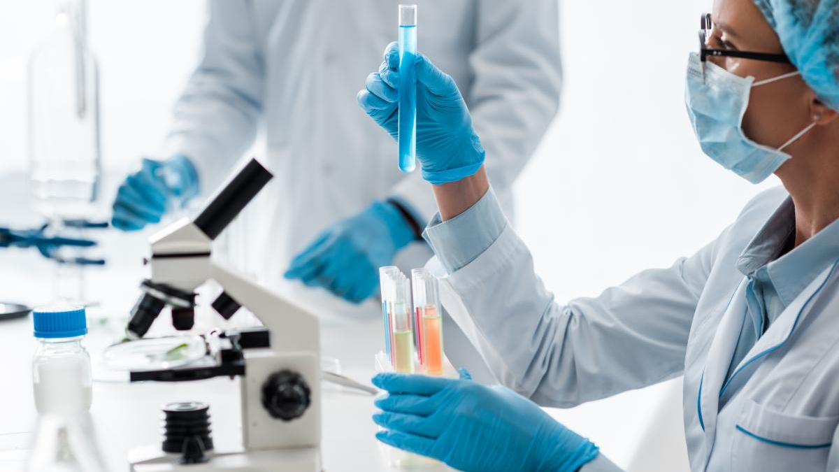 Lab technician in gloves and a mask holds a test tube with blue liquid, with a microscope and more tubes in the background.