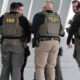 Group of officers in tactical vests labeled POLICE ICE, standing in a circle for briefing.
